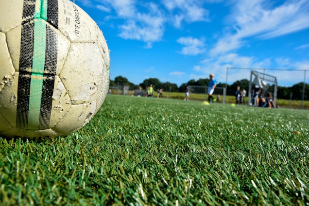 A well-used soccer ball lies on a lush green grass playing field under a bright blue sky with scattered clouds. In the background, a group of children in sportswear is visible, some standing and some sitting near a goalpost. The scene is situated outdoors, with a sense of motion and activity in a sports setting.