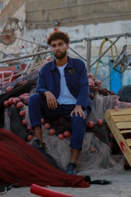 A person with curly hair and a beard wearing a blue jacket and pants sits on a pile of fishing nets. Around the nets, there are red floats. The background features industrial equipment and a building with peeling paint and exposed pipes.