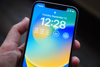 A person holds a smartphone displaying a brightly colored lock screen. The screen shows the time as 12:28 on Tuesday, September 13, along with multiple world clock times for different cities. The phone's interface includes a battery indicator and signal status at the top.