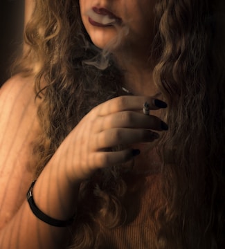A person with long wavy hair holds a cigarette between their fingers, with smoke drifting from their mouth. The dim lighting casts soft shadows, creating a serene and contemplative atmosphere.