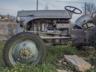 An old, weathered tractor sits in a grassy area, with visible signs of rust and wear on the metal parts. The engine and components are exposed, showing intricate details and a patina from age. A large wheel with a faded, rusty hub is prominently featured in the foreground. The tractor is set against a background of a wire fence and a concrete wall, with a partly visible rural landscape.