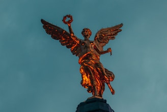 A golden statue of an angel with outstretched wings stands majestically against a backdrop of a teal sky. The angel holds a laurel wreath in one hand and appears to be illuminated by warm light, highlighting its intricate details.