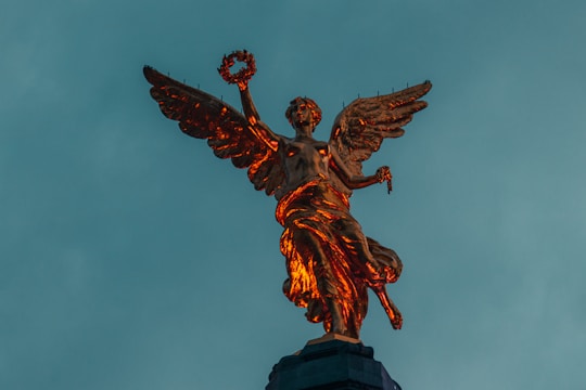 A golden statue of an angel with outstretched wings stands majestically against a backdrop of a teal sky. The angel holds a laurel wreath in one hand and appears to be illuminated by warm light, highlighting its intricate details.