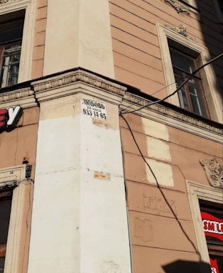 A street corner of a beige building with decorative architectural elements and two windows. A sign with black text and a phone number is posted on the corner. Part of a logo with a red background is visible on the right side of the image.