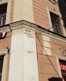 A street corner of a beige building with decorative architectural elements and two windows. A sign with black text and a phone number is posted on the corner. Part of a logo with a red background is visible on the right side of the image.