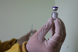 A gloved hand holds a vial of Pfizer-BioNTech vaccine with a blurred background, creating a clinical and focused scene.