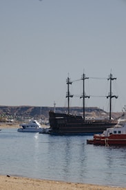 A large, dark-colored ship with masts resembling a pirate ship is situated in a calm harbor. Surrounding it are smaller modern boats and yachts. The background features rocky hills and a clear blue sky. The foreground shows a sandy shore touching the water.