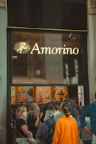 A group of people queue outside a store with large windows displaying colorful posters of gelato. The storefront has a prominent sign with the name 'Amorino' above it. The atmosphere seems busy with various individuals waiting.