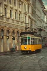 A vintage yellow tram travels along a cobblestone street lined with historical European buildings. The architecture features ornate details with arches and large windows. Two pedestrians walk on the sidewalk beside a row of Fendi storefronts.