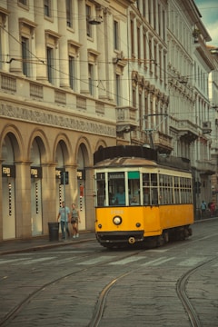 A vintage yellow tram travels along a cobblestone street lined with historical European buildings. The architecture features ornate details with arches and large windows. Two pedestrians walk on the sidewalk beside a row of Fendi storefronts.