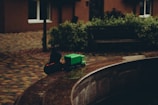A young child in a dark hoodie is playing with a bright green toy truck near a stone ledge. The setting appears to be an outdoor area with cobblestone paving and a bench surrounded by greenery.