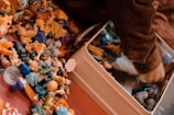 A collection of small, colorful plastic toys is scattered on a surface next to a child’s arm, which is reaching into a metal container. The toys include various shapes and characters in bright colors like yellow, blue, and orange. The child appears to be wearing a brown, ribbed sweater.