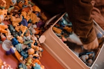 A collection of small, colorful plastic toys is scattered on a surface next to a child’s arm, which is reaching into a metal container. The toys include various shapes and characters in bright colors like yellow, blue, and orange. The child appears to be wearing a brown, ribbed sweater.