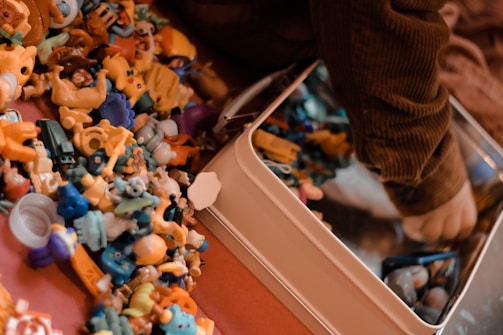 A collection of small, colorful plastic toys is scattered on a surface next to a child’s arm, which is reaching into a metal container. The toys include various shapes and characters in bright colors like yellow, blue, and orange. The child appears to be wearing a brown, ribbed sweater.