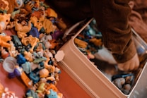 A collection of small, colorful plastic toys is scattered on a surface next to a child’s arm, which is reaching into a metal container. The toys include various shapes and characters in bright colors like yellow, blue, and orange. The child appears to be wearing a brown, ribbed sweater.