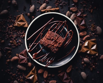 A rich, indulgent chocolate dessert is presented on a dark plate, drizzled with melted chocolate. Surrounding the dessert, several pieces of chocolate and cocoa beans are artfully scattered on a dark wooden surface, enhancing the image's decadent and luxurious feel.