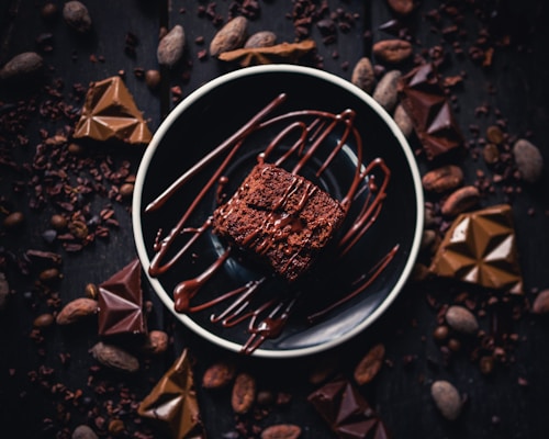 A rich, indulgent chocolate dessert is presented on a dark plate, drizzled with melted chocolate. Surrounding the dessert, several pieces of chocolate and cocoa beans are artfully scattered on a dark wooden surface, enhancing the image's decadent and luxurious feel.