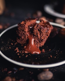 A rich, molten chocolate cake sits on a dark plate, with a smooth, glossy flow of chocolate lava oozing from the center. The texture of the cake is moist and crumbly, adding depth to its appearance, and the background is softly out of focus, emphasizing the dessert.