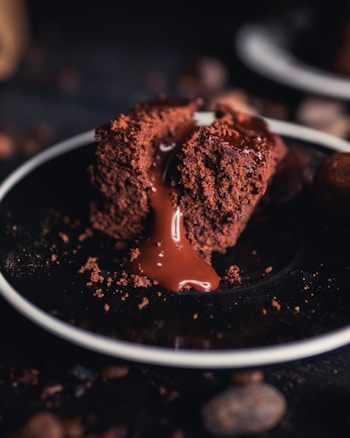 A rich, molten chocolate cake sits on a dark plate, with a smooth, glossy flow of chocolate lava oozing from the center. The texture of the cake is moist and crumbly, adding depth to its appearance, and the background is softly out of focus, emphasizing the dessert.