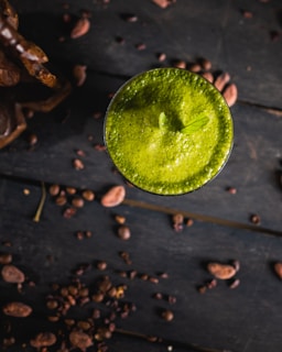 A vibrant green smoothie or drink in a glass is positioned in the center on a dark wooden table. Surrounding the glass are scattered cocoa beans and pieces creating a rustic and organic atmosphere. A small, fresh leaf garnishes the top of the drink.