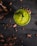 A vibrant green smoothie or drink in a glass is positioned in the center on a dark wooden table. Surrounding the glass are scattered cocoa beans and pieces creating a rustic and organic atmosphere. A small, fresh leaf garnishes the top of the drink.