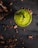 A vibrant green smoothie or drink in a glass is positioned in the center on a dark wooden table. Surrounding the glass are scattered cocoa beans and pieces creating a rustic and organic atmosphere. A small, fresh leaf garnishes the top of the drink.