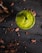 A vibrant green smoothie or drink in a glass is positioned in the center on a dark wooden table. Surrounding the glass are scattered cocoa beans and pieces creating a rustic and organic atmosphere. A small, fresh leaf garnishes the top of the drink.