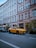 A yellow luxury car is parked along a street in front of a building with multiple windows and a storefront named Pickarnia. There are other cars parked nearby, and small trees or shrubs line the sidewalk. The scene appears to be in an urban setting with a clear blue sky above.