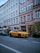 A yellow luxury car is parked along a street in front of a building with multiple windows and a storefront named Pickarnia. There are other cars parked nearby, and small trees or shrubs line the sidewalk. The scene appears to be in an urban setting with a clear blue sky above.
