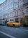 A yellow luxury car is parked along a street in front of a building with multiple windows and a storefront named Pickarnia. There are other cars parked nearby, and small trees or shrubs line the sidewalk. The scene appears to be in an urban setting with a clear blue sky above.
