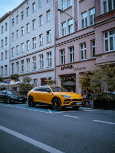 A yellow luxury car is parked along a street in front of a building with multiple windows and a storefront named Pickarnia. There are other cars parked nearby, and small trees or shrubs line the sidewalk. The scene appears to be in an urban setting with a clear blue sky above.