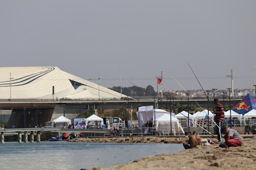 A waterfront area with people engaged in fishing. In the background, an extensive, modern architectural structure with wavy lines is visible. Multiple white tents are set up along the water, and a flag with a red background is on display. The overall ambiance suggests a recreational or event space.