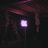 Neon sign displaying the words 'Good Vibes' in bright pink, suspended in a dimly lit outdoor setting with wooden beams and hanging plants.