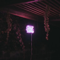 Neon sign displaying the words 'Good Vibes' in bright pink, suspended in a dimly lit outdoor setting with wooden beams and hanging plants.