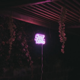 Neon sign displaying the words 'Good Vibes' in bright pink, suspended in a dimly lit outdoor setting with wooden beams and hanging plants.