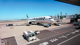 A large Finnair airplane is parked at an airport gate with jet bridges extended. Ground service vehicles, including a tug, are positioned around the aircraft, and workers are seen around the tarmac. The sky is clear and blue, contributing to the busy yet orderly environment of the airport.