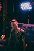 A young man wearing a floral shirt stands outdoors at night. Above him is a neon sign reading 'Good Vibes' in blue light. To his side, a heart-shaped lamp glows softly, adding a warm hue to the scene. String lights and floral decorations surround the area, creating a relaxed and festive atmosphere.