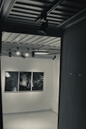 A dimly lit gallery room featuring three abstract black and white artworks displayed on a white wall. The ceiling has several spotlights focused on the artwork, casting subtle shadows in the room.
