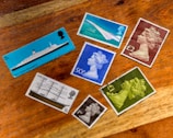 A collection of seven vintage postage stamps displayed on a wooden surface. The stamps feature various designs including a ship labeled 'RMS Queen Elizabeth 2', and images of the Concorde airplane. Several stamps depict a profile of a monarch wearing a crown in different colors such as blue, green, and brown. One stamp shows a ship named 'Cutty Sark'. All stamps are positioned in a scattered arrangement, showing a mix of vibrant and muted colors.