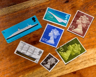 A collection of seven vintage postage stamps displayed on a wooden surface. The stamps feature various designs including a ship labeled 'RMS Queen Elizabeth 2', and images of the Concorde airplane. Several stamps depict a profile of a monarch wearing a crown in different colors such as blue, green, and brown. One stamp shows a ship named 'Cutty Sark'. All stamps are positioned in a scattered arrangement, showing a mix of vibrant and muted colors.