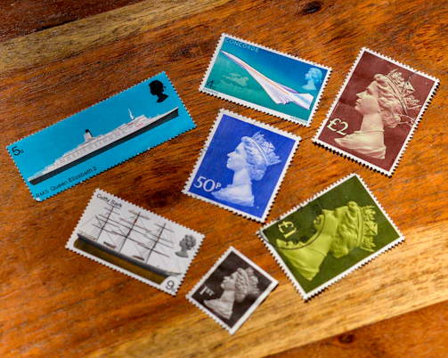 A collection of seven vintage postage stamps displayed on a wooden surface. The stamps feature various designs including a ship labeled 'RMS Queen Elizabeth 2', and images of the Concorde airplane. Several stamps depict a profile of a monarch wearing a crown in different colors such as blue, green, and brown. One stamp shows a ship named 'Cutty Sark'. All stamps are positioned in a scattered arrangement, showing a mix of vibrant and muted colors.