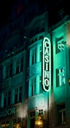 A vibrant neon sign displaying the word 'CASINO' is prominently illuminated against the facade of a building, showcasing intricate architectural details and a classic design. The sign emits a bright teal glow, casting light on the surrounding area and creating a lively atmosphere at night.