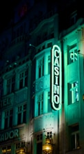 A vibrant neon sign displaying the word 'CASINO' is prominently illuminated against the facade of a building, showcasing intricate architectural details and a classic design. The sign emits a bright teal glow, casting light on the surrounding area and creating a lively atmosphere at night.