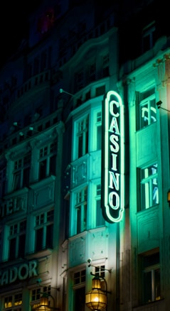 A vibrant neon sign displaying the word 'CASINO' is prominently illuminated against the facade of a building, showcasing intricate architectural details and a classic design. The sign emits a bright teal glow, casting light on the surrounding area and creating a lively atmosphere at night.
