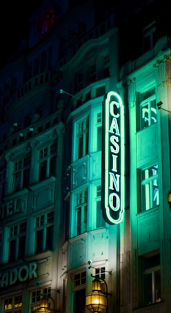 A vibrant neon sign displaying the word 'CASINO' is prominently illuminated against the facade of a building, showcasing intricate architectural details and a classic design. The sign emits a bright teal glow, casting light on the surrounding area and creating a lively atmosphere at night.