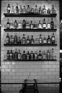 A monochrome image featuring several shelves stocked with various bottles of liquor against a backdrop of white subway tiles. The bottles are neatly arranged and occupy multiple levels. The overall setting suggests a bar or a stylized retail display.