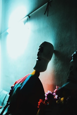 A dimly lit scene featuring a stylized skull figure with a vibrant, colorful attire. The figure casts a shadow on the wall, and the light from above creates a dramatic contrast. There are flowers at the base contributing to the decorative setup.