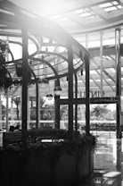 A black and white photograph of a modern indoor space with high ceilings and large windows. The architecture features metal and glass structures. There is a rounded wooden kiosk adorned with hanging plants and pendant lights. Outside, trees and foliage can be seen through the windows, along with a sign that appears to read 'PAQUETE'. Reflections are visible on the shiny floor.