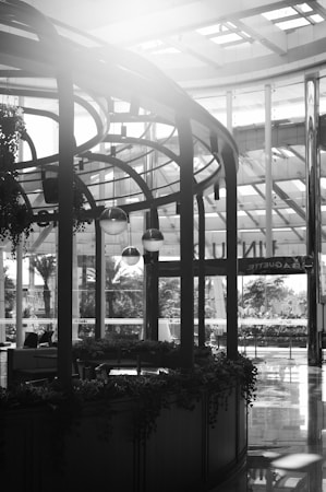 A black and white photograph of a modern indoor space with high ceilings and large windows. The architecture features metal and glass structures. There is a rounded wooden kiosk adorned with hanging plants and pendant lights. Outside, trees and foliage can be seen through the windows, along with a sign that appears to read 'PAQUETE'. Reflections are visible on the shiny floor.