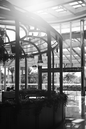A black and white photograph of a modern indoor space with high ceilings and large windows. The architecture features metal and glass structures. There is a rounded wooden kiosk adorned with hanging plants and pendant lights. Outside, trees and foliage can be seen through the windows, along with a sign that appears to read 'PAQUETE'. Reflections are visible on the shiny floor.